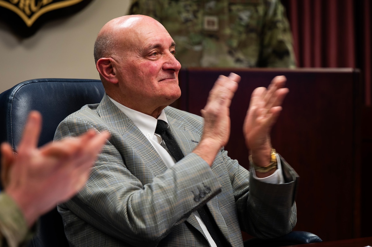 White man in grey suit sits and claps.