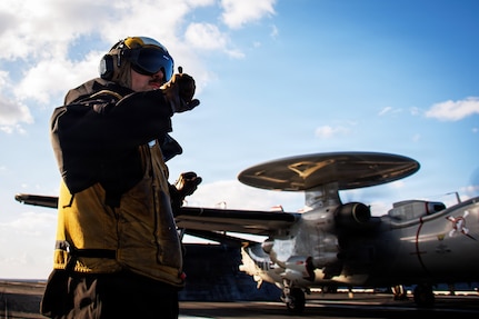 A sailor wearing a flight suit, gloves and head, ear and eye protection signals with his hands on the deck of a ship during daytime, with an aircraft in the background.
