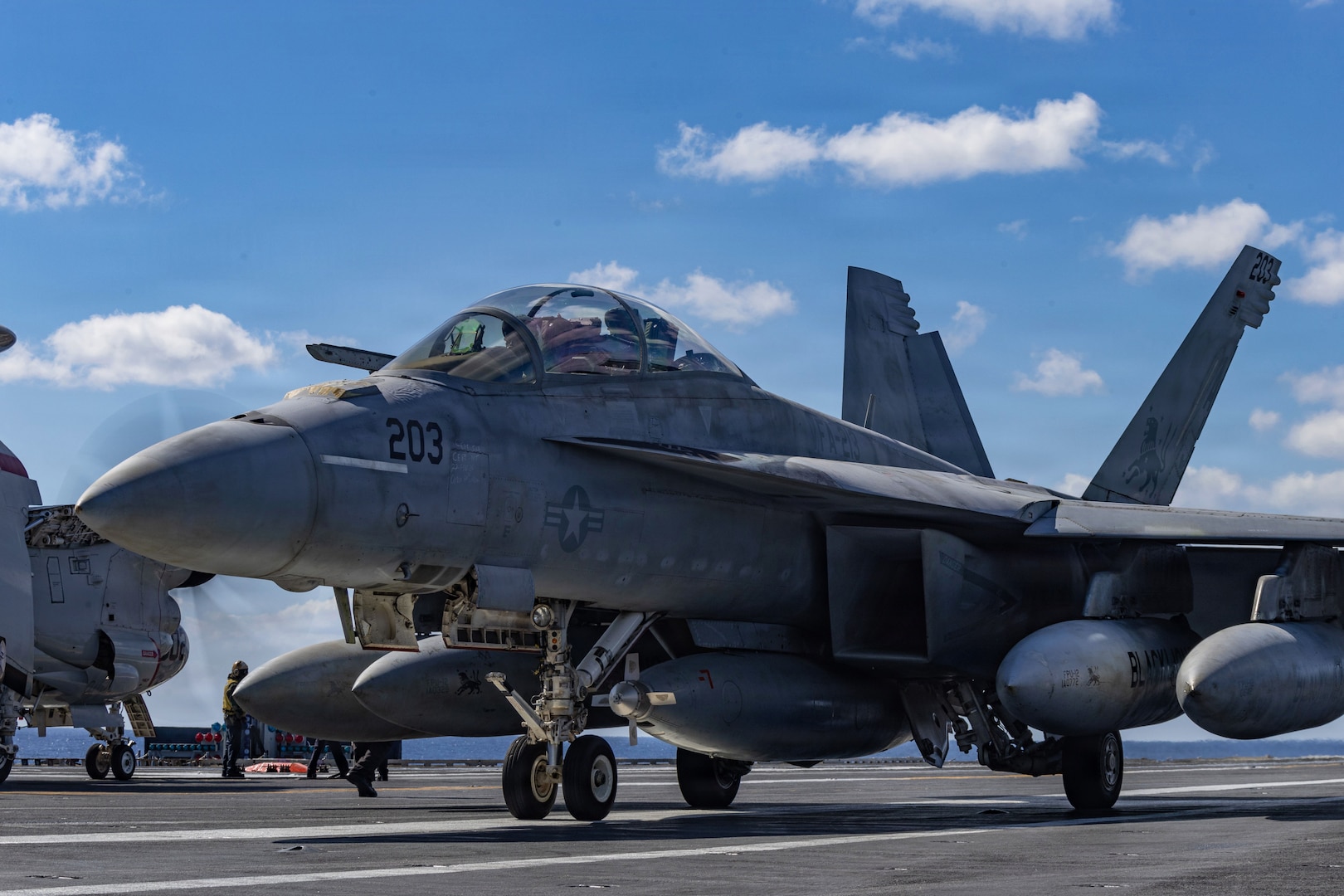 A jet is parked on the deck of a ship during daytime, with another jet and people standing in the background.