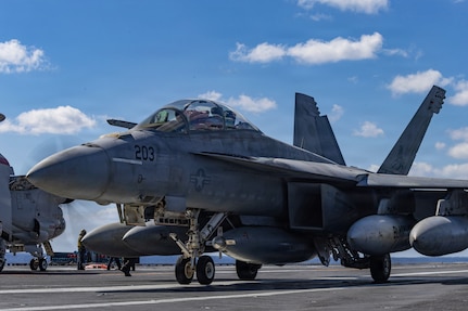 A jet is parked on the deck of a ship during daytime, with another jet and people standing in the background.