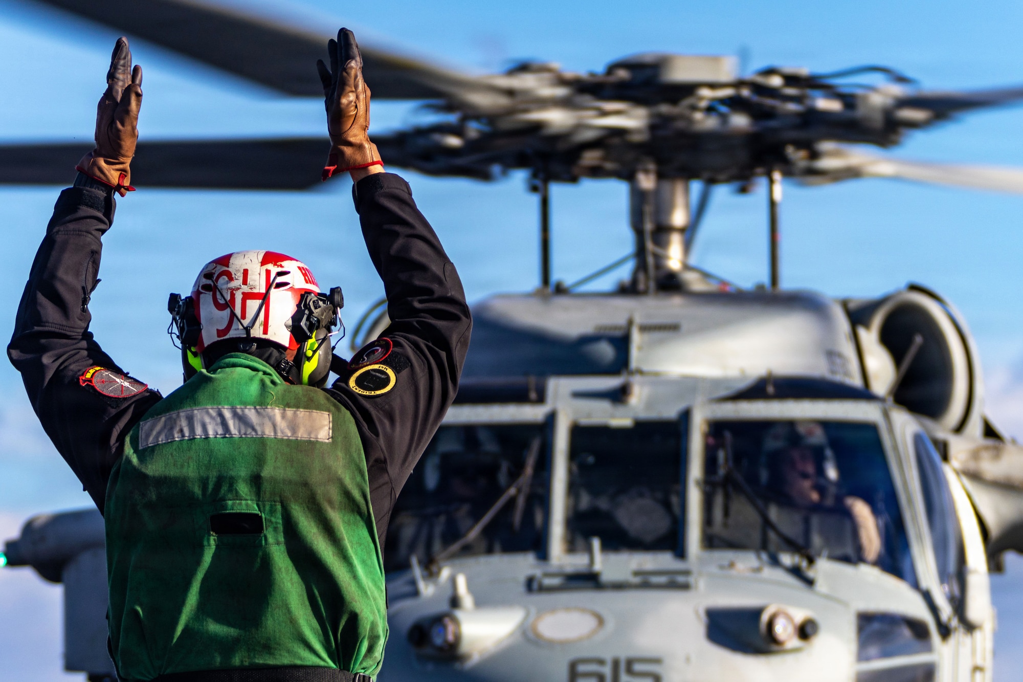 A sailor wearing a flight suit, gloves, a helmet and a green vest raises both arms while facing a helicopter during daytime, with a blue sky in the background.