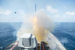 A missile launches from a ship in an open body of water during daytime, emitting smoke and orange flames.
