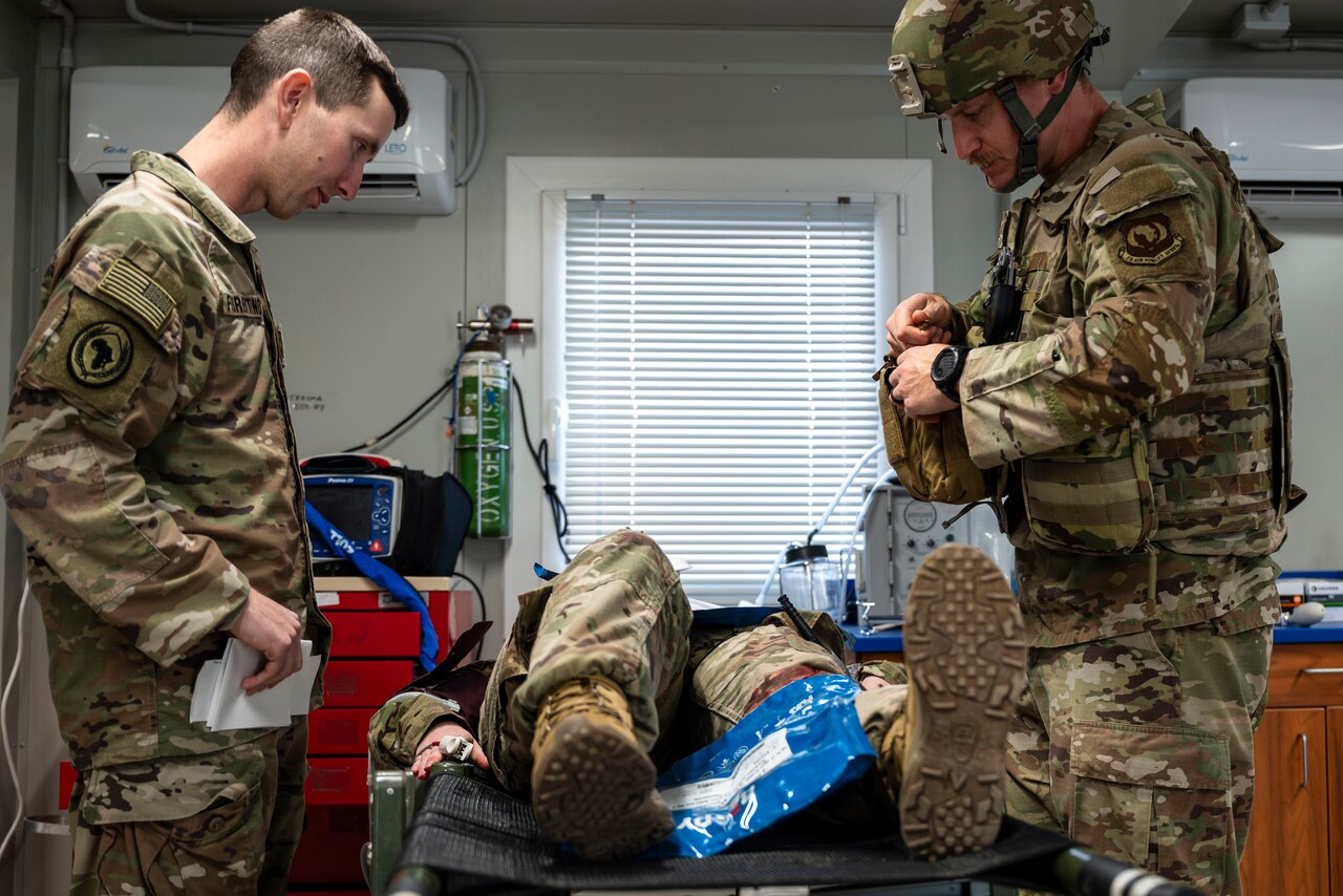 Two soldiers look down at a soldier lying on a medical bed.