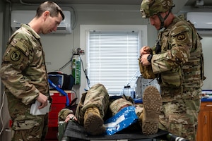 Two soldiers look down at a soldier lying on a medical bed.