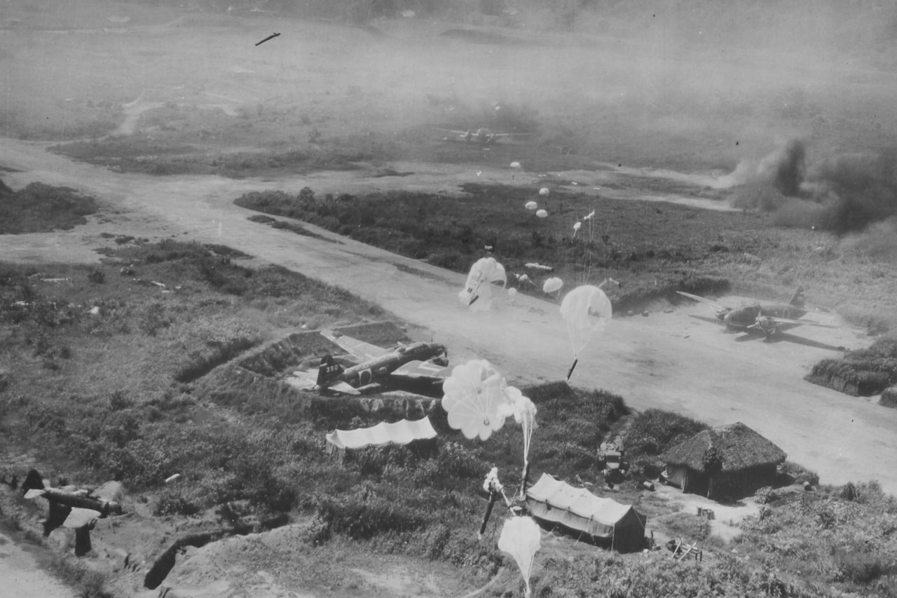 An aerial view of an island airstrip as parachutes drop on it.