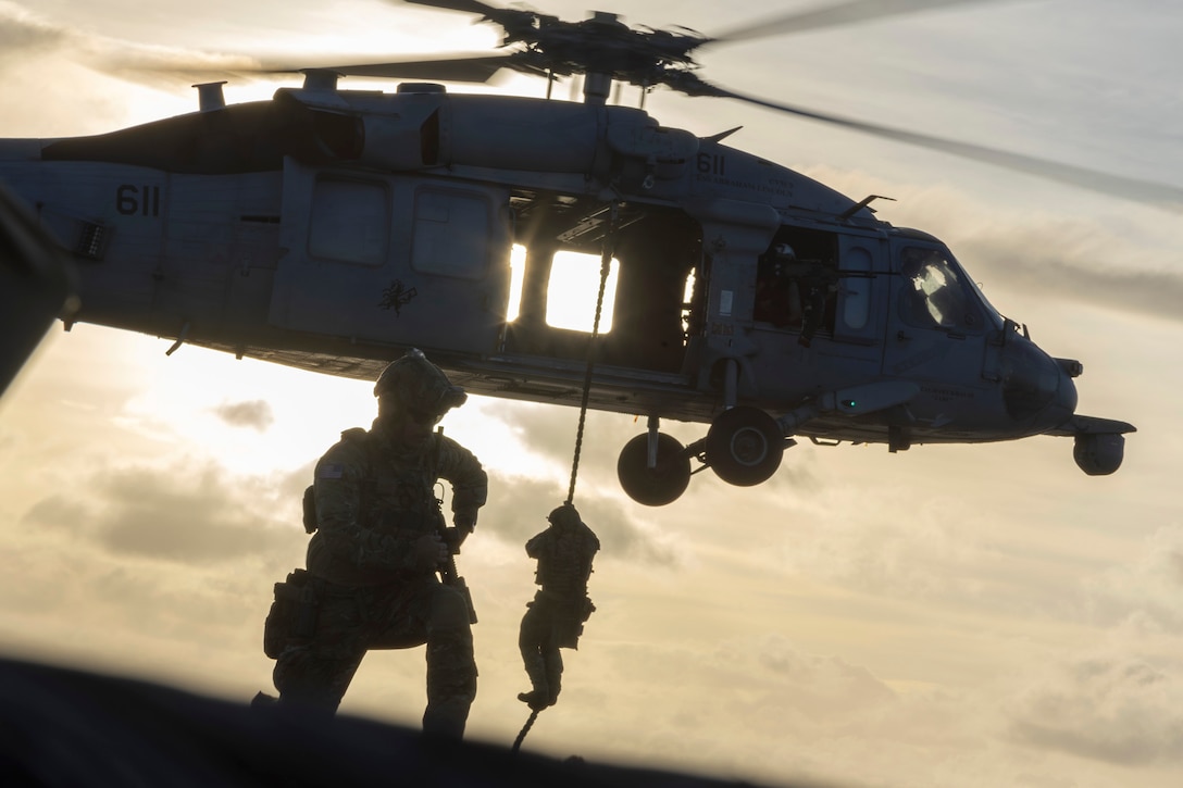A sailor fast-ropes from a hovering helicopter onto a ship at sea as a fellow sailor watches as shown in silhouette against a sunlit sky.