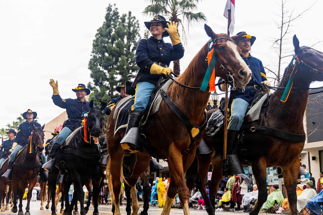 Soldiers riding horses on a street wave to crowd of parade watchers.