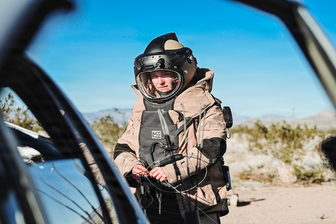A sailor in protective gear works with wire near a partially visible vehicle against a blue sky with mountains in the background.