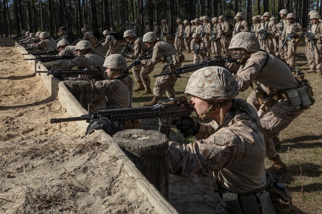 Marine Corps recruits kneel behind a wooden structure to fire weapons in a wooded area.
