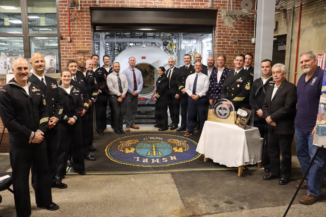 GROTON, Conn. (Nov. 6, 2025) Past and present members of Naval Submarine Medical Research Laboratory (NSMRL) gather to cut a ceremonial ribbon following the certification ceremony for the Genesis Chamber. NSMRL, part of Navy Medicine Research & Development and based out of Groton, Connecticut, sustains the readiness and superiority of U.S. undersea warfighters through innovative health and performance research and works to lead the world in delivering science solutions to ensure undersea warrior dominance. (U.S. Navy photo by Emily Swedlund /Released)
