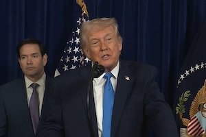 A person in a business suit speaks into a microphone at a lectern. Another person in similar attire stands in the background next to an American flag.