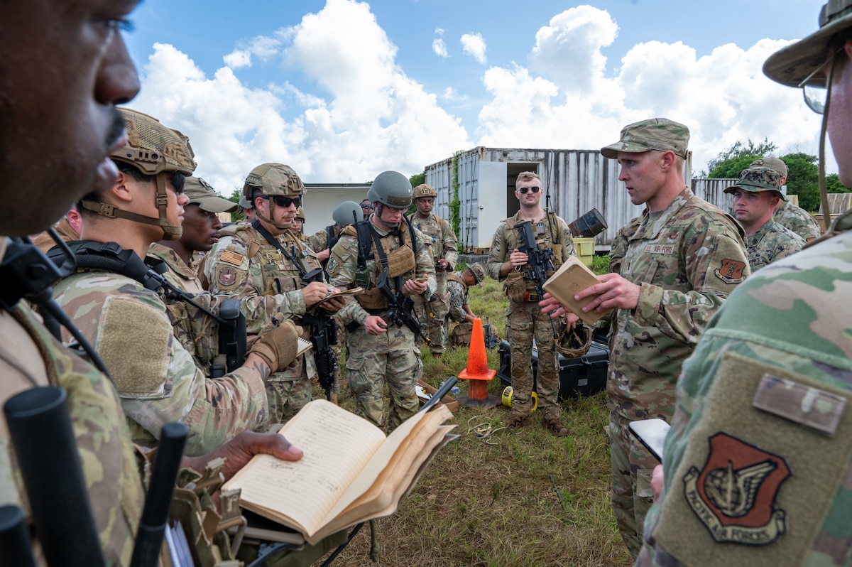 U.S. Airmen and U.S. Marines in military fatigues form a circle outside around a military leader during a mission briefing.