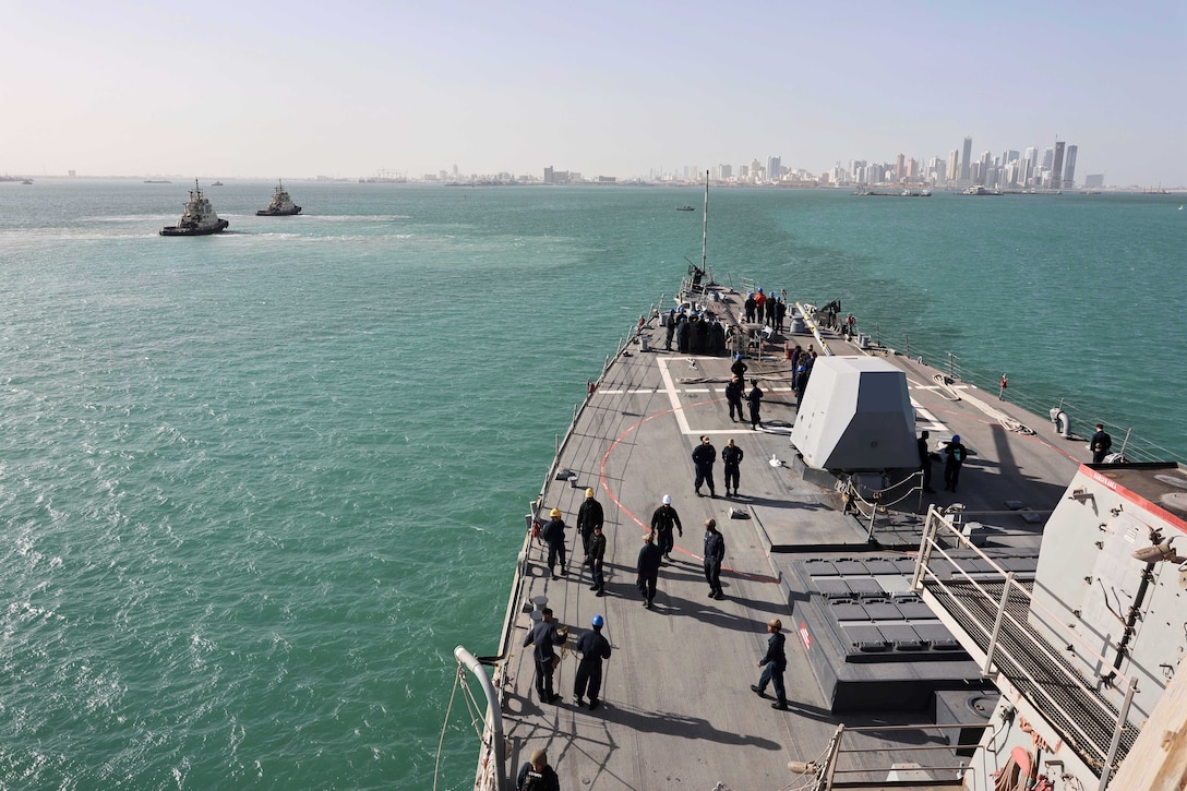 Sailors work on a ship while sailing next to other boats toward a skyline during the day.
