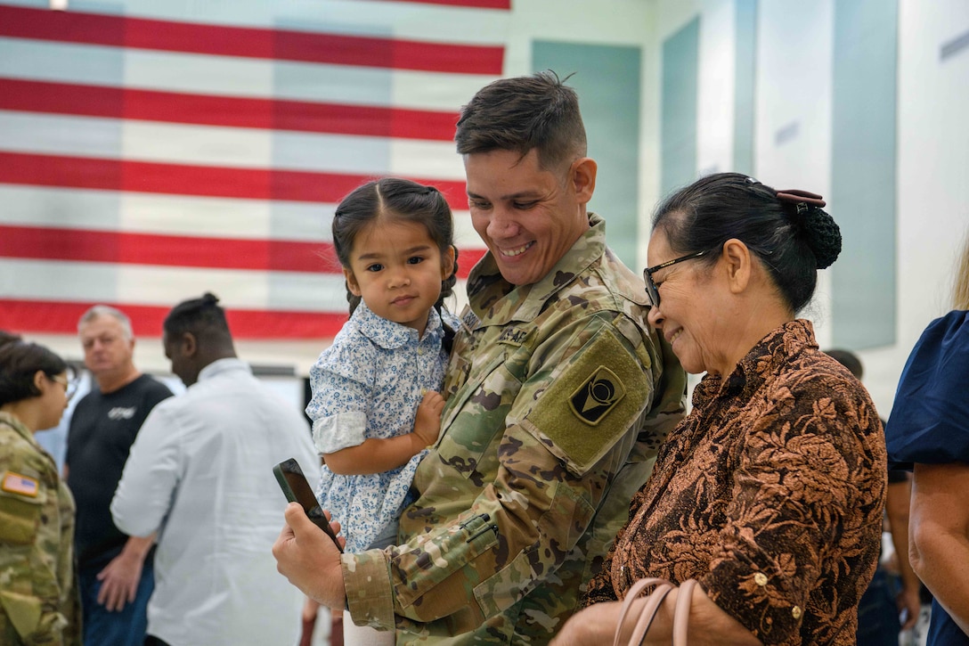 A soldier holding a child smiles for a selfie next to a loved one as an American flag hangs on the left.