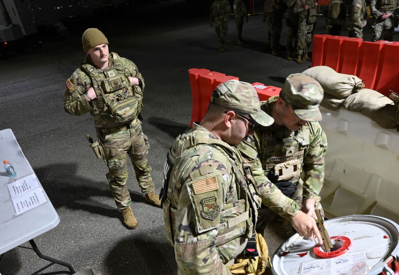 A man wearing a military camouflage uniform clears is pistol into a white drum while two other men dressed in similar attire watch.