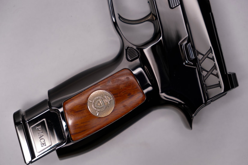 Image of a shiny pistol with wood on the stock, the word PEACE on the end of the magazine, and the letters XXI inscribed on it near the hammer.
