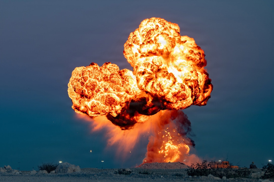 An explosion creates a large fireball in a dark, blue sky in a desert-like area.