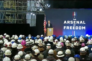 Secretary of War Pete Hegseth stands at a podium while speaking to a crowd of people wearing white helmets.