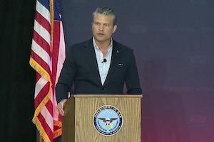 A person in a business suit is speaking from behind a podium with an American flag in the background.