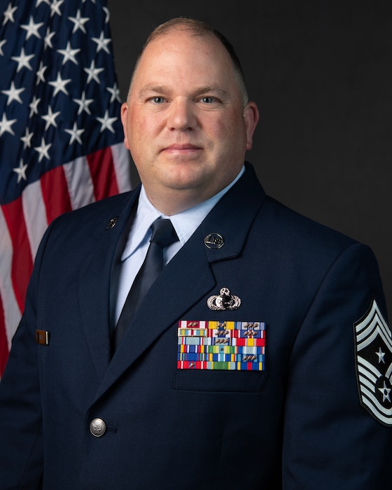 An Air National Guard command chief master sergeant poses for a studio portrait with the U.S. flag over his right shoulder. He is wearing service dress blues.