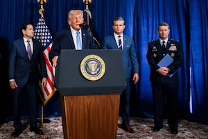 President Trump speaks into a microphone at a lectern. Three other people in similar attire stand in the background next to two flags.