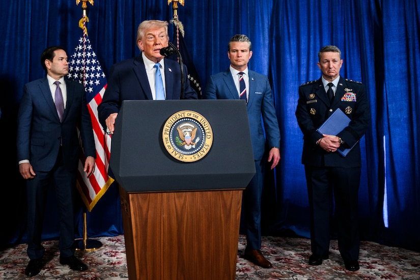 A person in a business suit speaks into a microphone at a lectern. Three other people in similar attire stand in the background next to two flags.