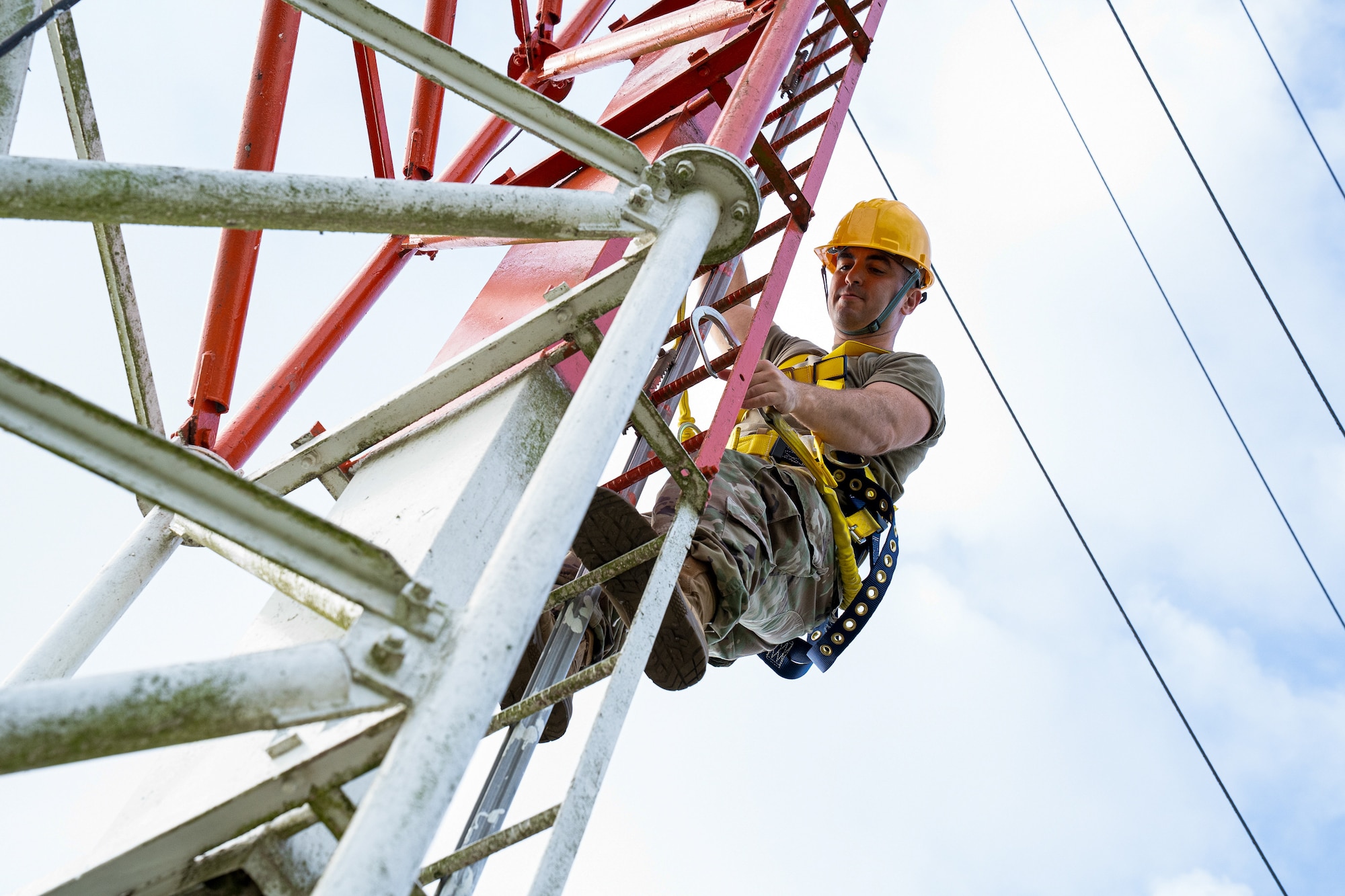 A serviceman, without his OCP top, climbs a ladder while wearing a yellow hard hat.