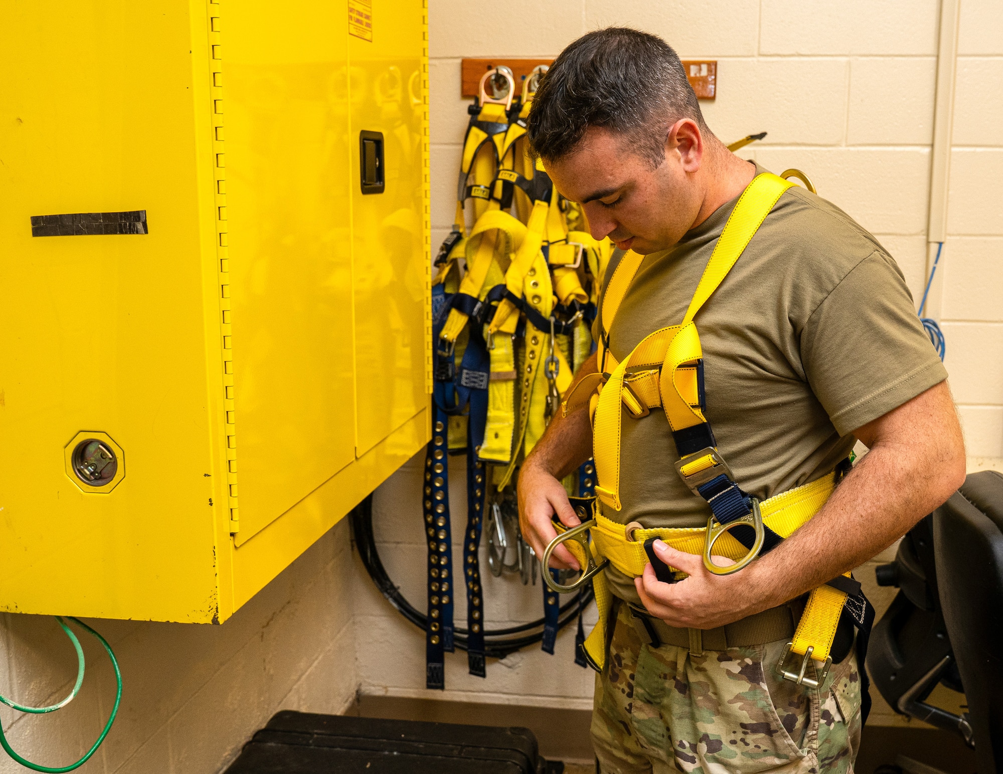 A serviceman, without his OCP top, adjusting a yellow climbing harness.