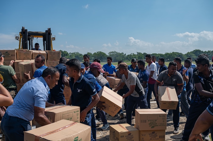 A crown of civilians organize carboard boxes around a large forklift.
