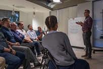 a speaker stands while talking to a group seated in chairs