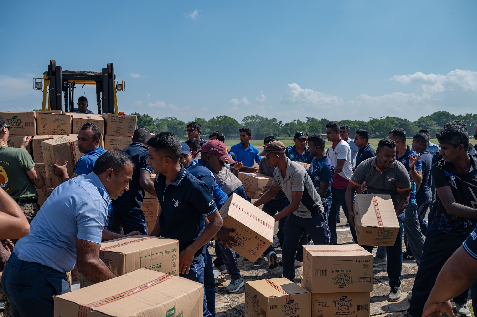 A crown of civilians organize carboard boxes around a large forklift.