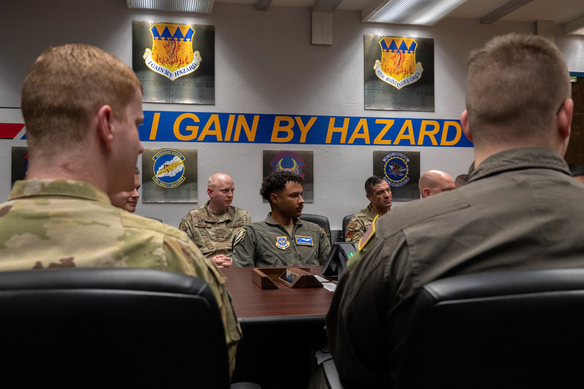 Airmen from the 317th Airlift Wing sit under their motto, "I gain by hazard," as they receive a holiday greeting call from Secretary of the Air Force Troy Meink at Dyess Air Force Base, Texas, Dec. 24, 2025. Meink praised the 317th AW for their exceptional service, a recent "highly effective" Foundational Readiness Inspection grade and completing 12 months of deployed operations in the Middle East. (U.S. Air Force photo by Airman William Neal)