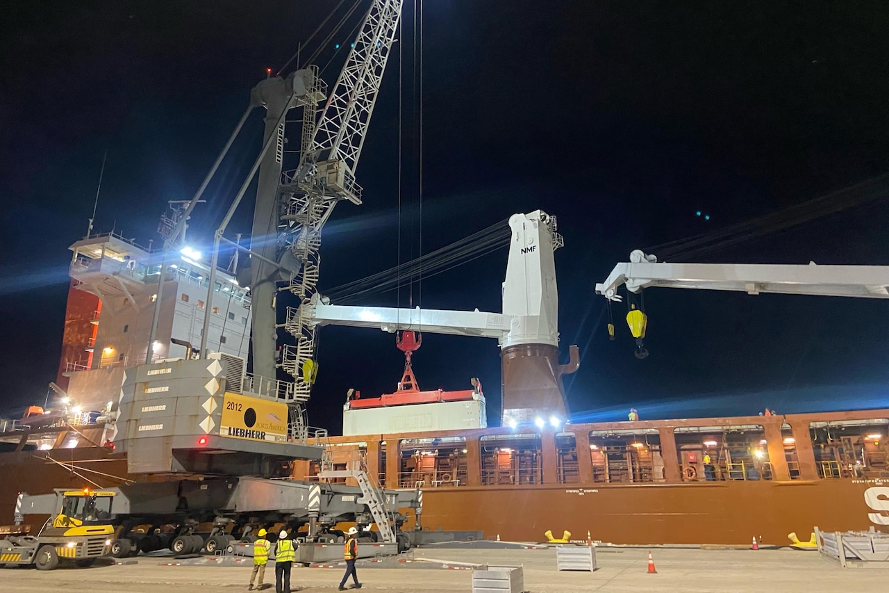 Three people wearing reflective vests and hard hats watch as a crane loads cargo onto a large docked ship.