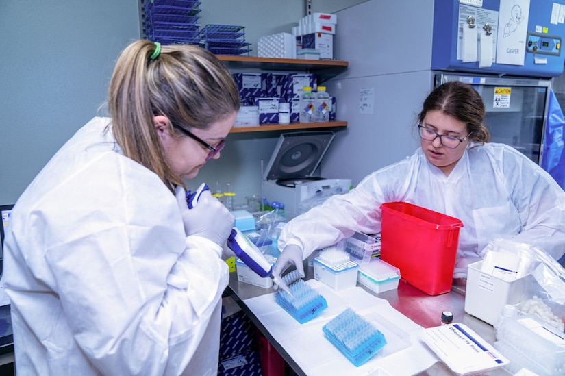 A woman in white coveralls and latex gloves uses laboratory equipment to fill small containers with blood as another woman in similar attire holds them.