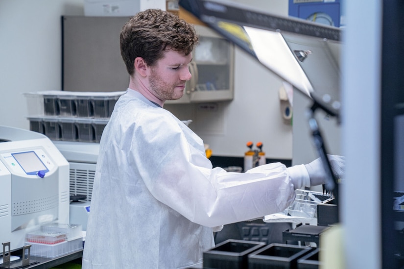 A man in white coveralls uses laboratory equipment to take DNA samples from small containers.