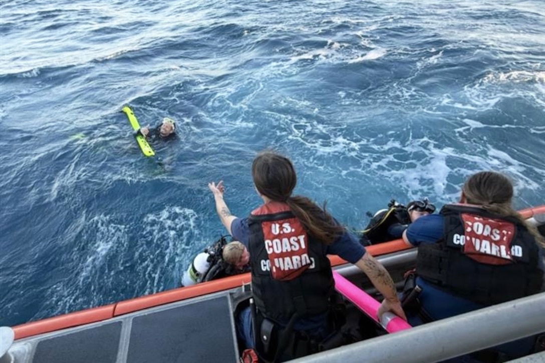 Coast Guardsmen wearing life vests aboard a boat extend their hands to help divers in a body of water.