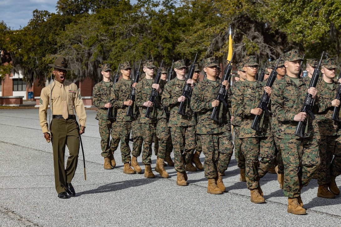 A Marine shouts to direct dozens of recruits marching in formation while carrying weapons.