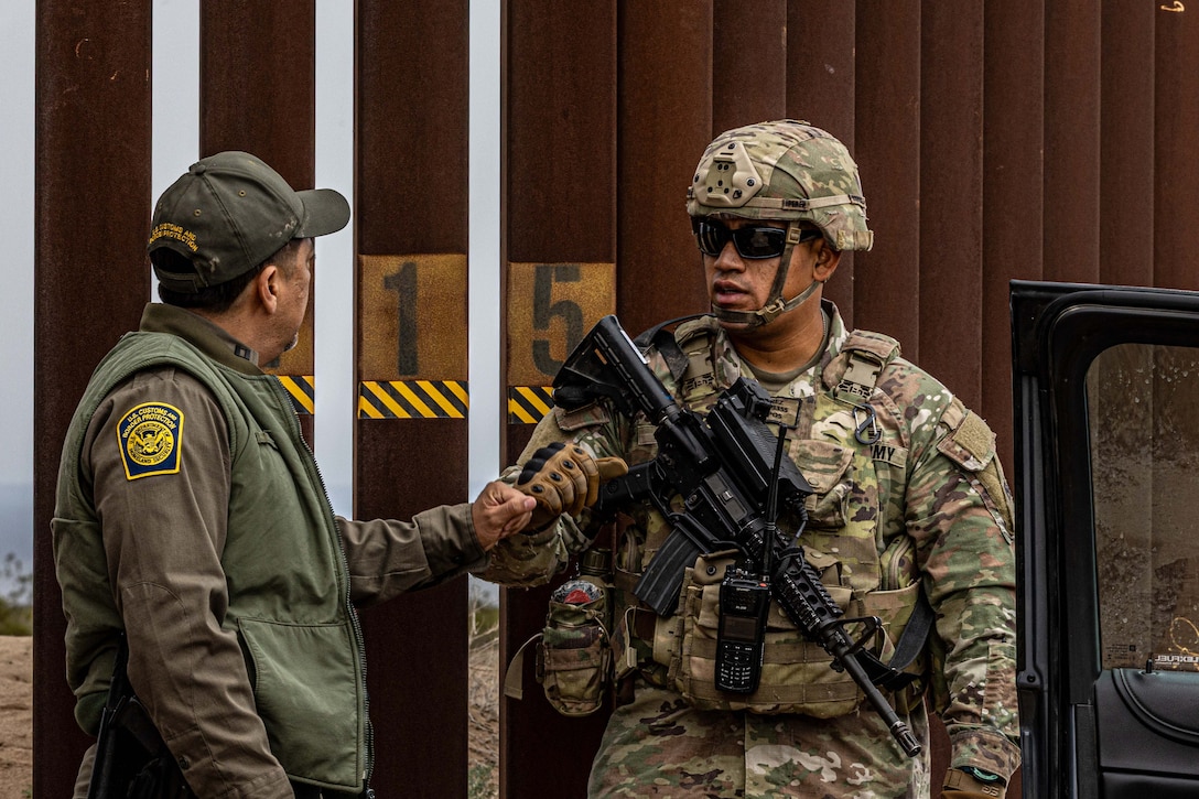 A soldier in tactical gear daps a U.S. Customs and Border Protection agent outside of a large, metal fence.