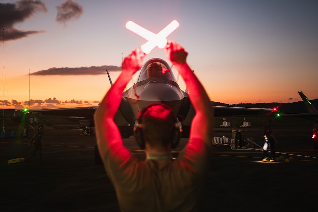 An airman uses red batons to signal to an aircraft as orangish hues are shown on the horizon.