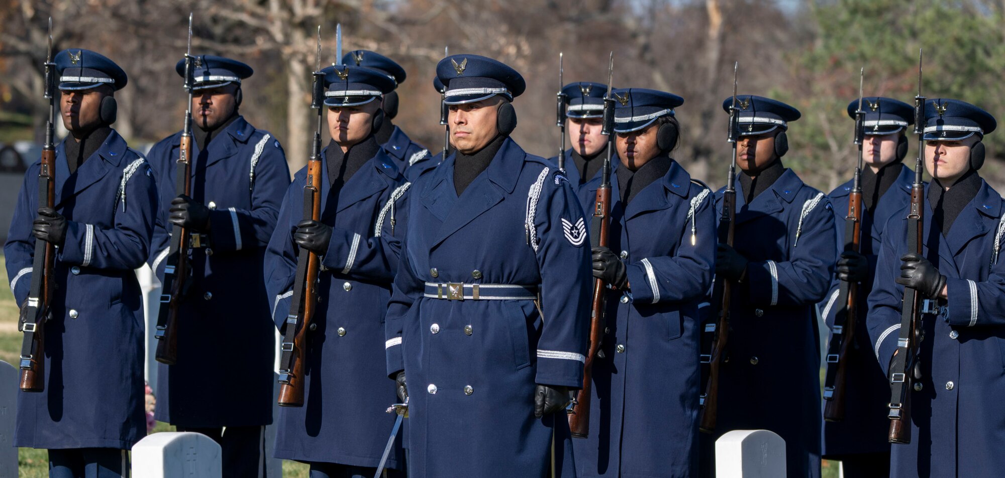An Honor Guardsman stands with a sword in front of ten Airmen standing with rifles.