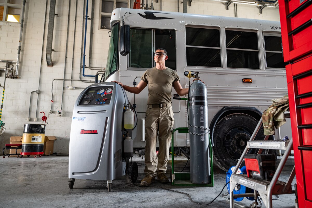 A technician standing next to a pressure cylinder and a box with gauges, and a bus in the background.