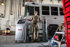A technician standing next to a pressure cylinder and a box with gauges, and a bus in the background.