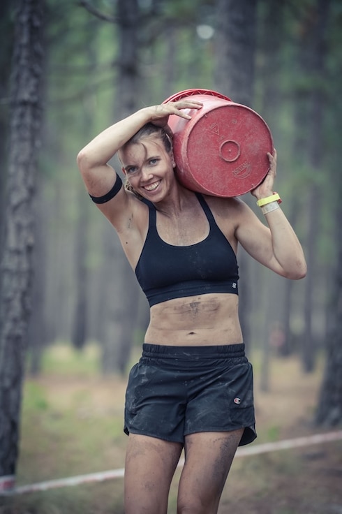 A girl covered in dirt smiles as she carries a weight over her shoulder