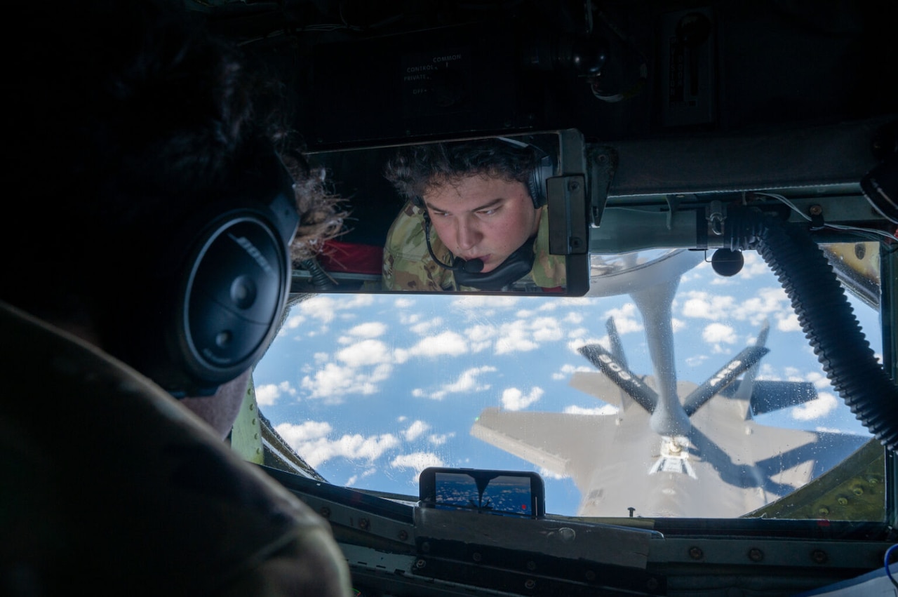 The reflection from an overhead mirror shows a man in a camouflage military uniform and headset looking out a window as a fuel boom connects to a military fighter jet flying below.