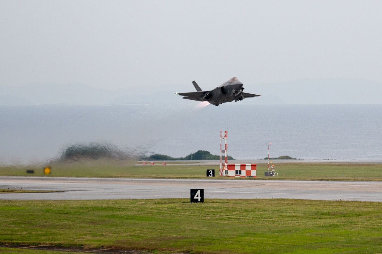 A military fighter jet takes off from a runway next to a large body of water, with mountains in the background.