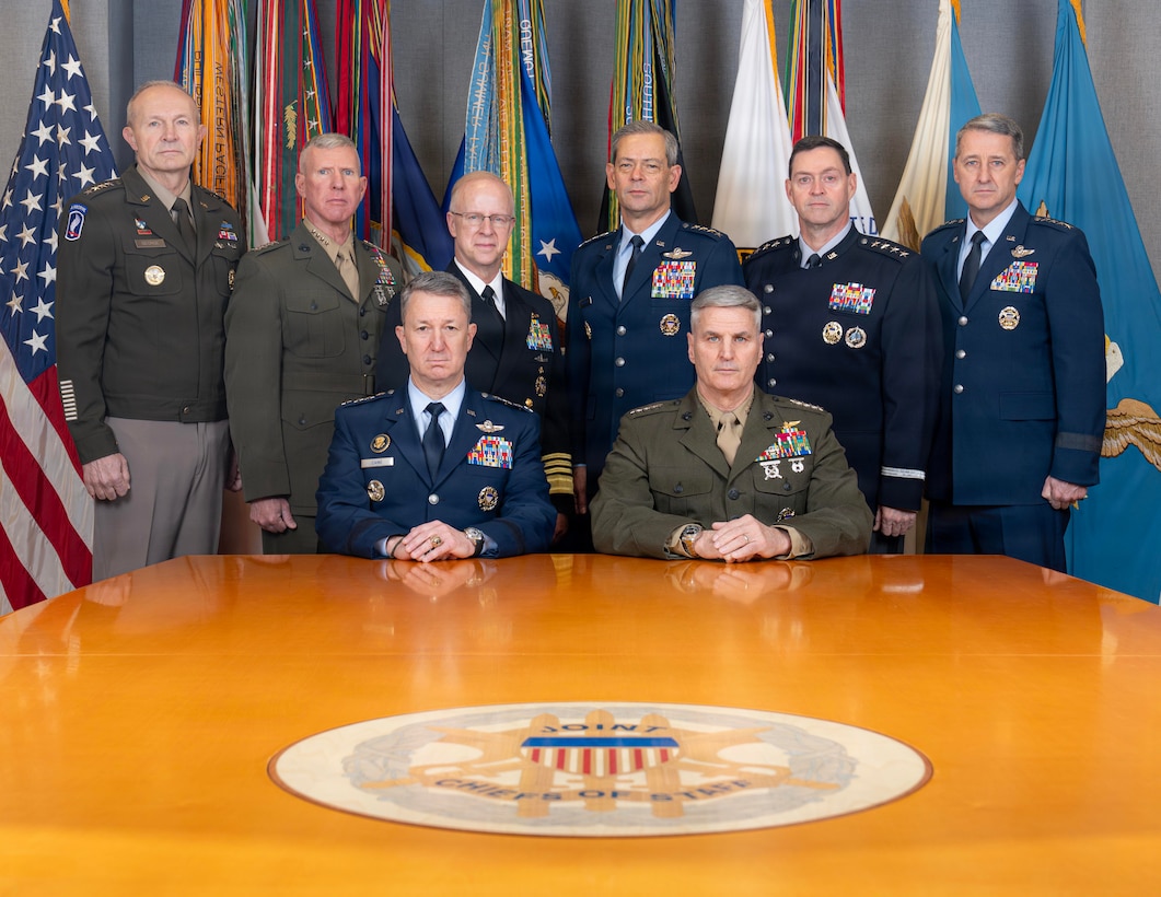 A group of eight men in uniform pose in front of standing flags and behind a large wooden table while two of them sit.