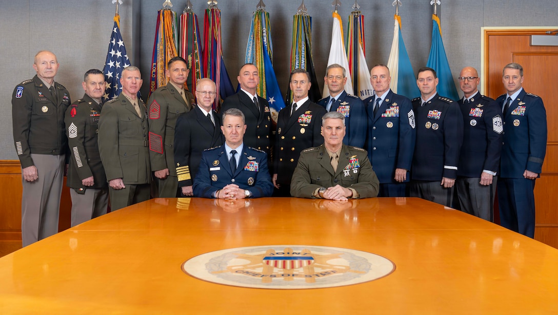 A group of 14 men in uniform pose in front of standing flags and behind a long wooden table while two are seated.