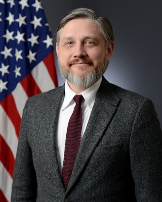 A person poses in a business suit in from of the American flag.