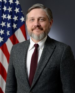 A person poses in a business suit in from of the American flag.