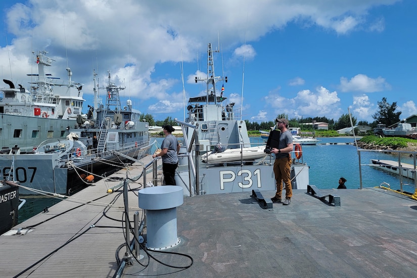 Two people stand on a dock near small military vessels.